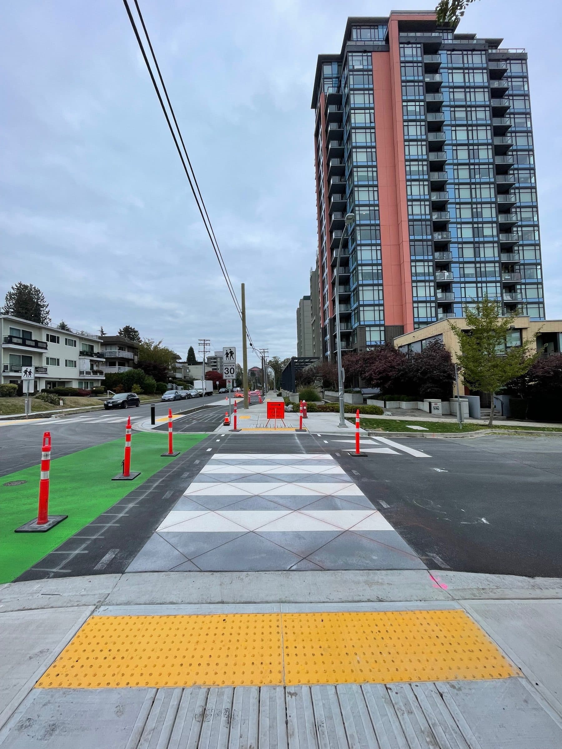 Three systems in one frame on the Agnes Greenway, New Westminster: a green coated bike lane, a white TrafficPatternsXD crossing and yellow tactile plates at the kerb, a tower beyond