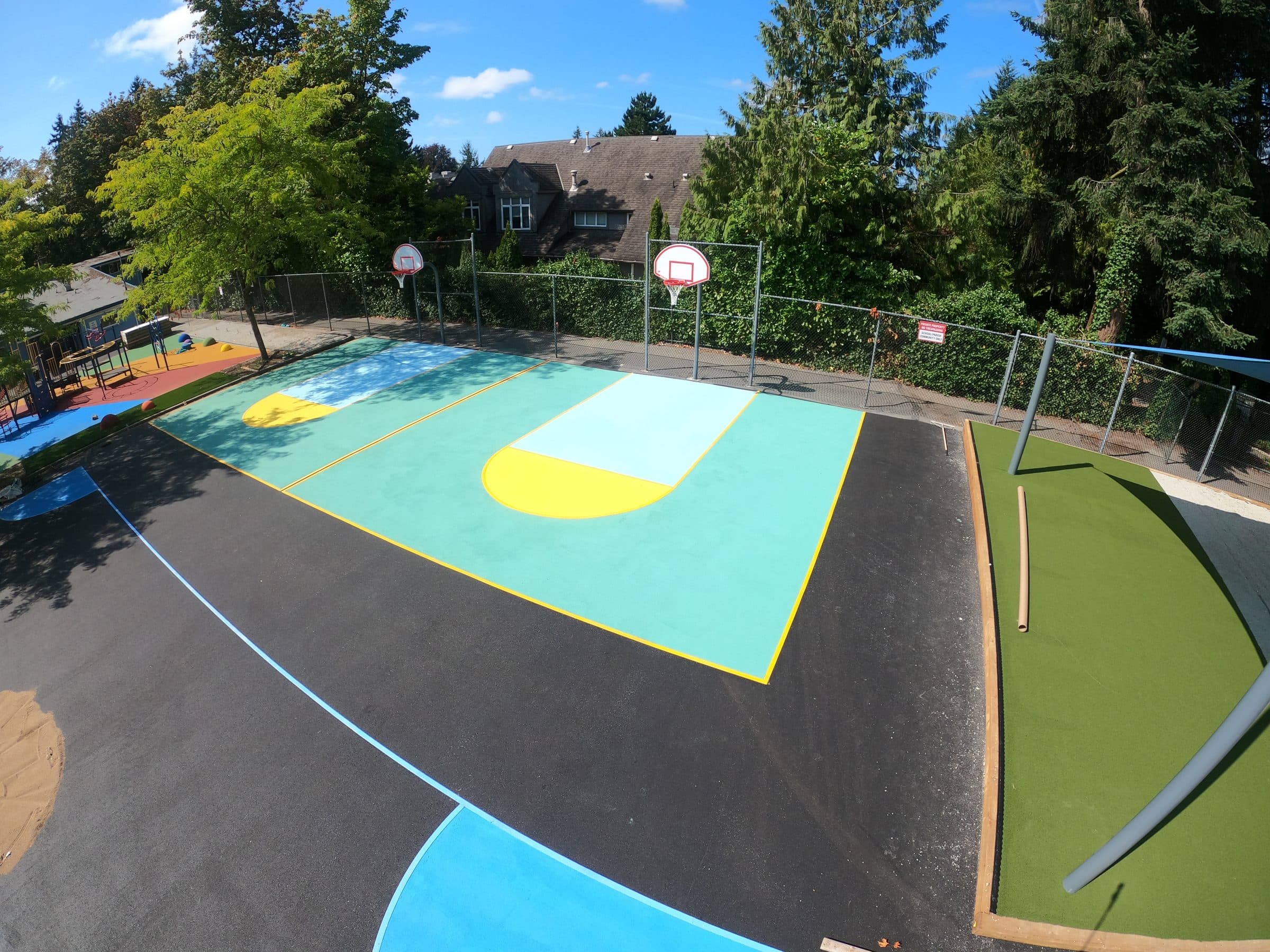 A teal and yellow StreetBond basketball court at a school in Vancouver, hoops and trees around it