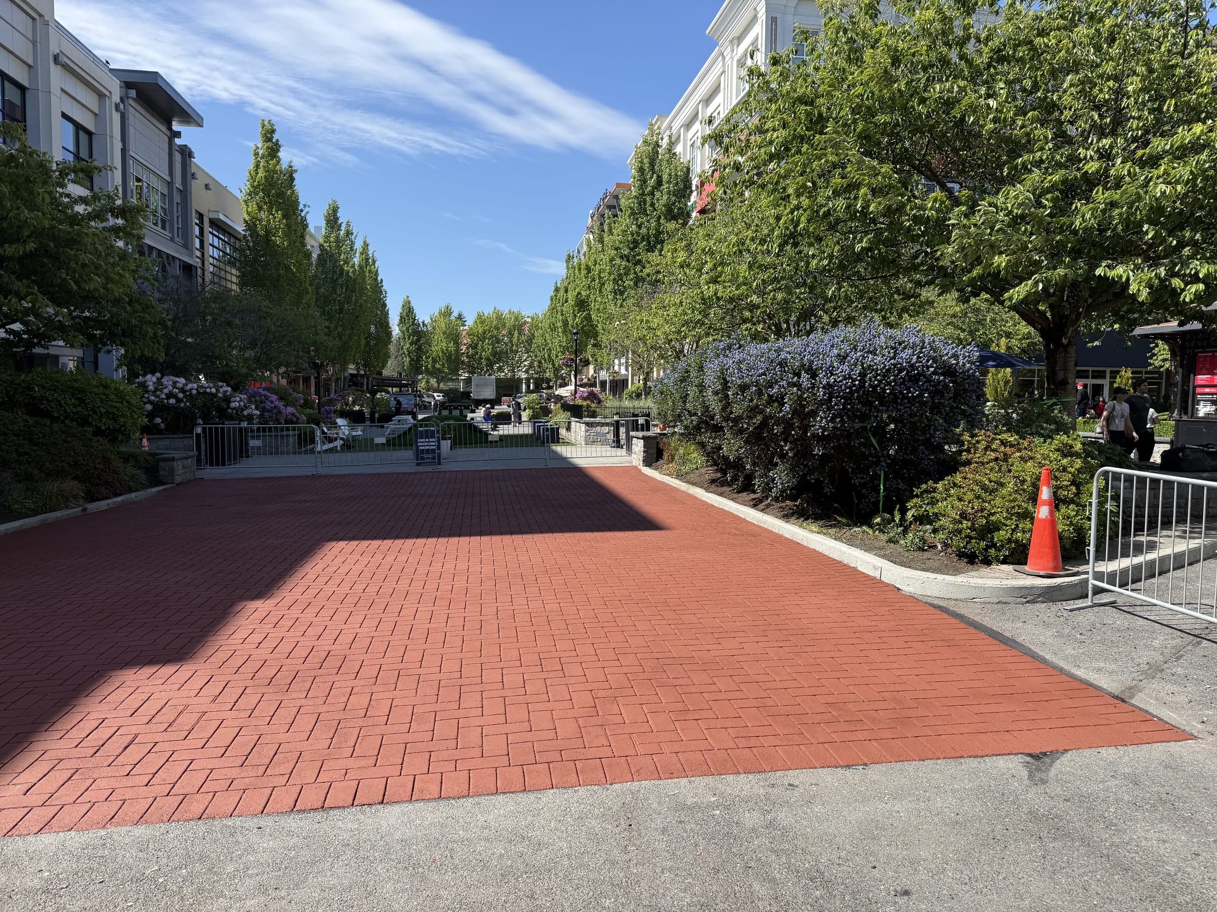 Brick-red StreetPrint stamped asphalt crossing at the edge of a town centre plaza in Victoria, with shops and trees beyond