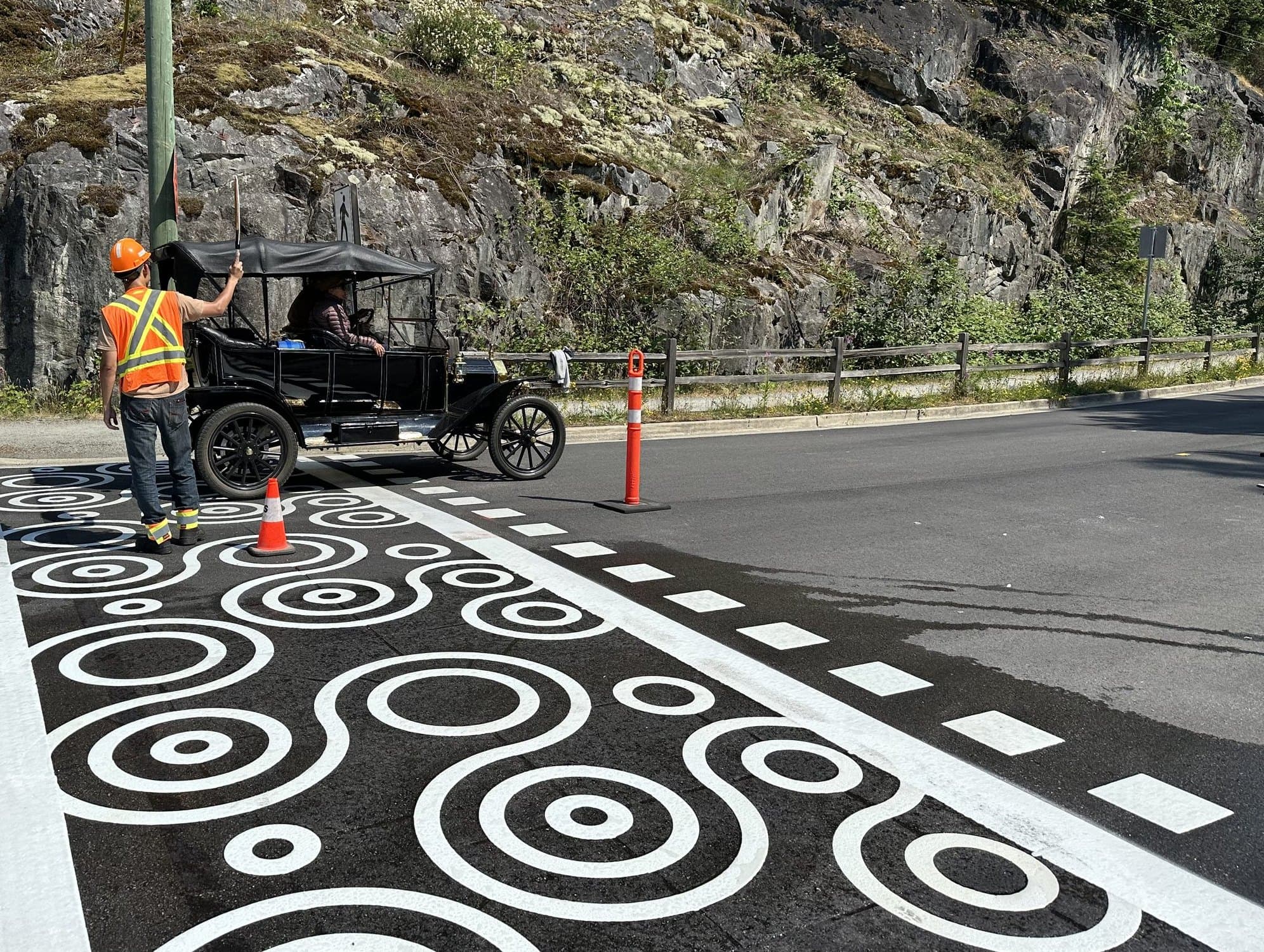 A Square One crew member in a hard hat and high-visibility vest waving a vintage car across a freshly coated StreetBond crossing on the Spirit Trail in West Vancouver