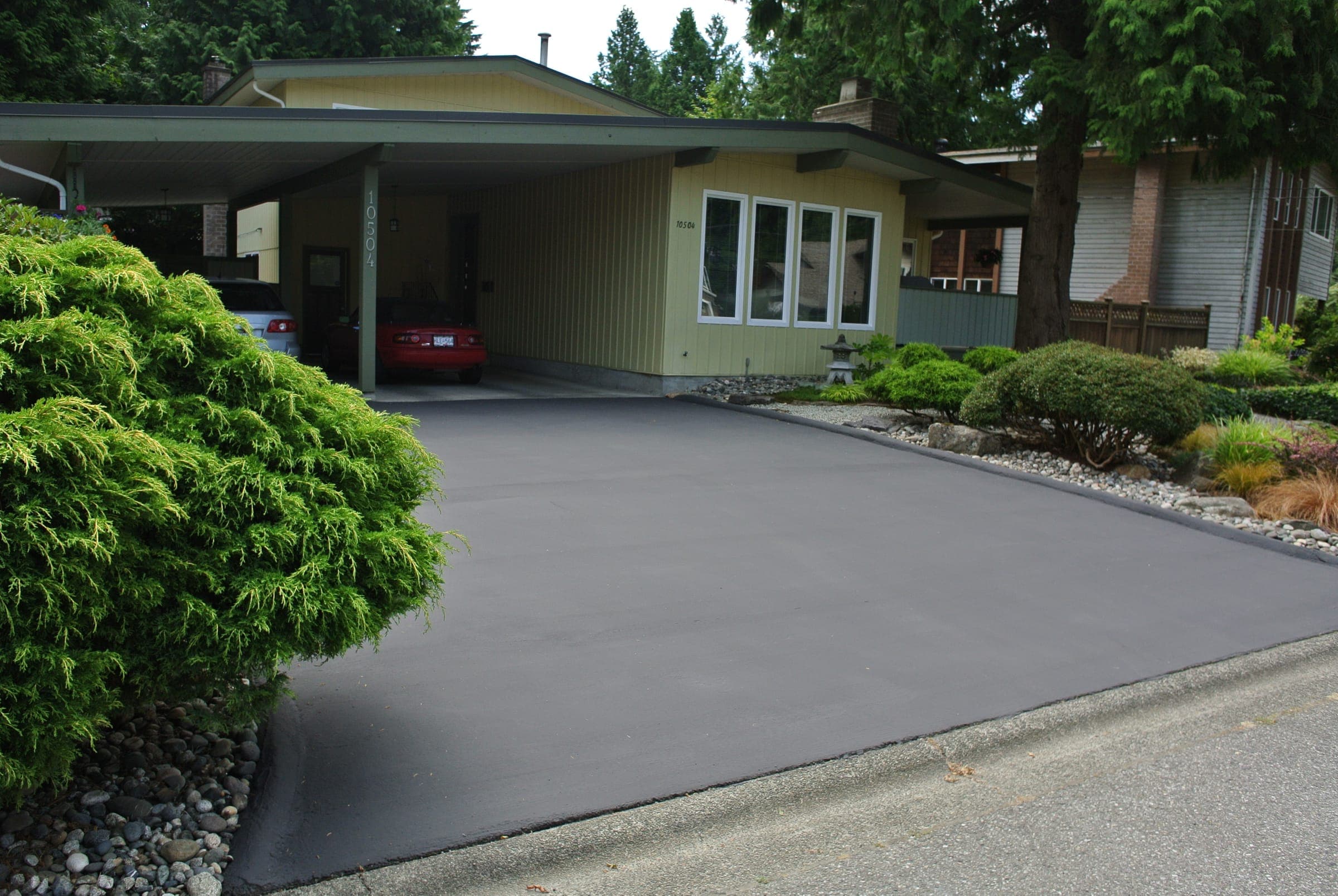 A residential driveway freshly coated black with DuraShield, running up to a carport beside a shrub garden